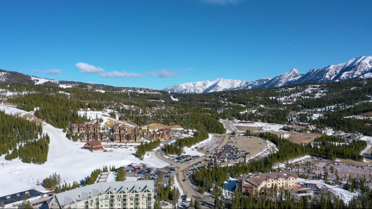 Aerial view reveals cars tightly packed into a snowy lot in Big Sky, Montana, framed by pine trees, rental condos, and towering alpine peaks just minutes from the nearby ski resort