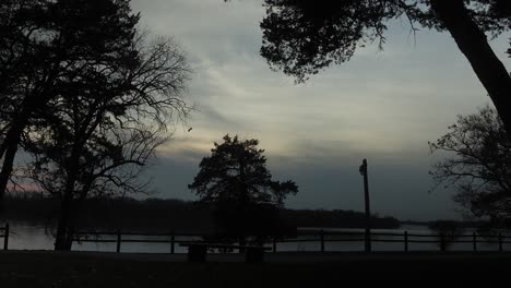 time lapse of a cloudy sunset over a large river with a wooden fence, trees, and a bench in the foreground