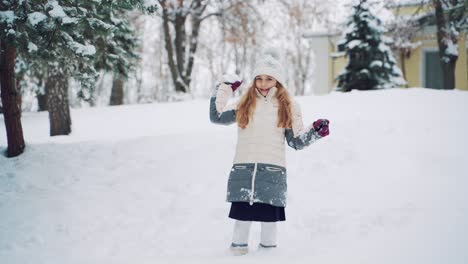 a beauty girl in a warm down jacket and hat holds a snowball in her hand and throws it forward on a cold weather in the winter in the hill near the house. slow motion