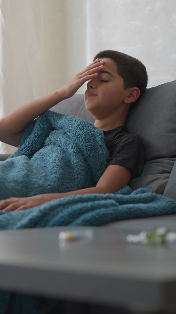 Young child suffering from flu cough lies on couch with thick blanket covering him, as he rests near window with curtain, with blurred medicine and tissue visible on table in foreground