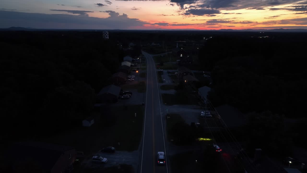 Peaceful night scene after sunset with driving car on street in calm neighborhood. Vehicle leaving home driveway driving on road. Aerial wide shot. Dusk scene in American Suburbia. Wide shot