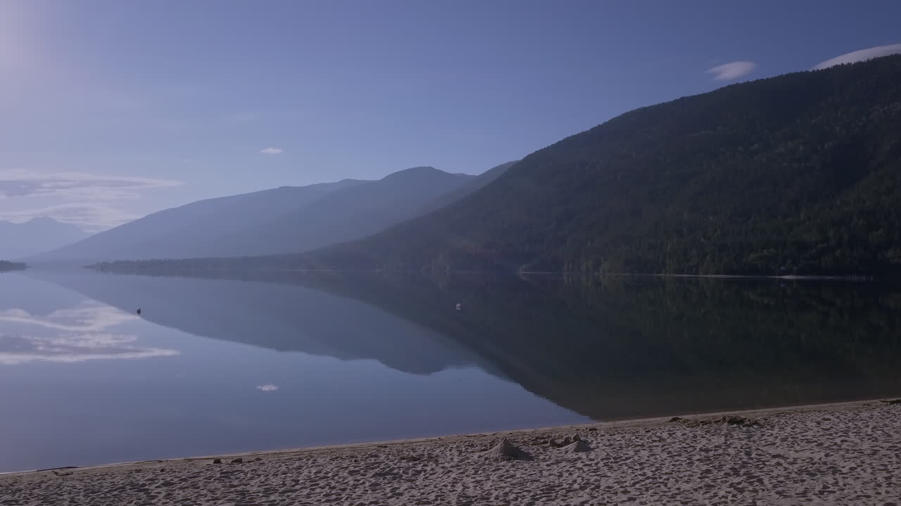 Calm summer morning over a lake with glass-like surface creating a nearly perfect reflection of the hills in the background. Blue sky with an early morning haze.