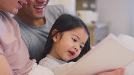 Familia-Con-Dos-Padres-En-La-Cama-En-Casa-Leyendo-Un-Cuento-A-Su-Hija