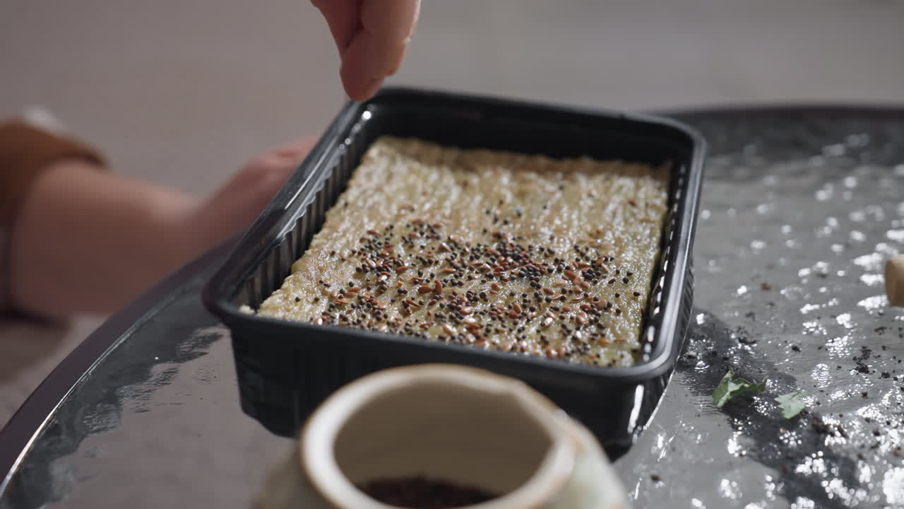 Female hands gently spreading tiny seeds from ceramic pot onto moist growing medium in black tray on glass table surrounded by gardening tools and plants, capturing detailed indoor sowing ritual