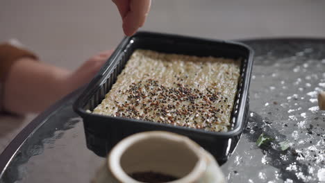 female hands gently spreading tiny seeds from ceramic pot onto moist growing medium in black tray on glass table surrounded by gardening tools and plants, capturing detailed indoor sowing ritual