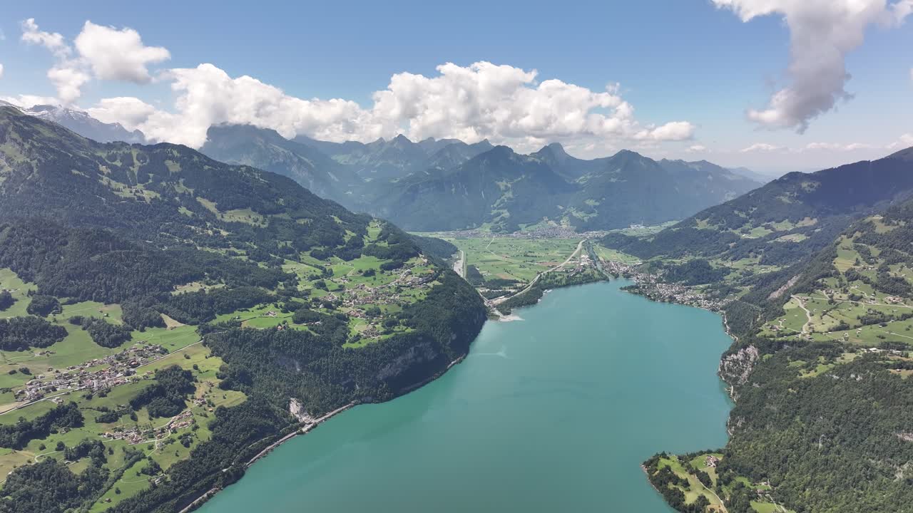 Panoramic aerial view of Walensee surrounded by green hills, alpine villages, and distant Swiss mountains under a bright summer sky