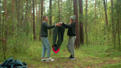 man and woman tourists bend down to fold tent fabric together in peaceful forest setting, surrounded by camping gear and tall trees, smiling warmly at each other