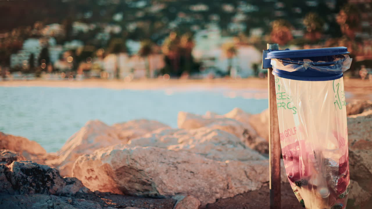 Transparent trash bag filled with waste hanging on a bin by rocky sea defences, with a blurred coastal town and beach in the background at golden hour