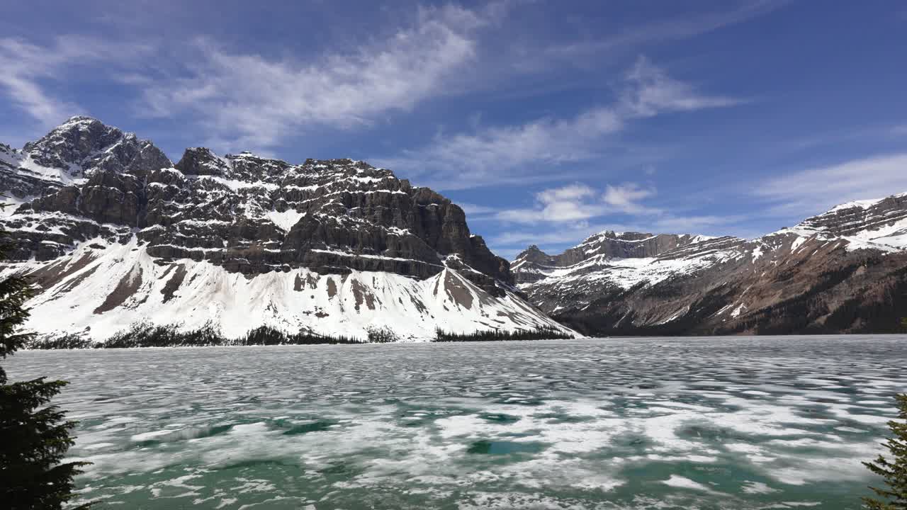 Lago Bow ghiacciato che mostra il ghiaccio che si scioglie, con montagne innevate e cielo blu nel Parco Nazionale di Banff.