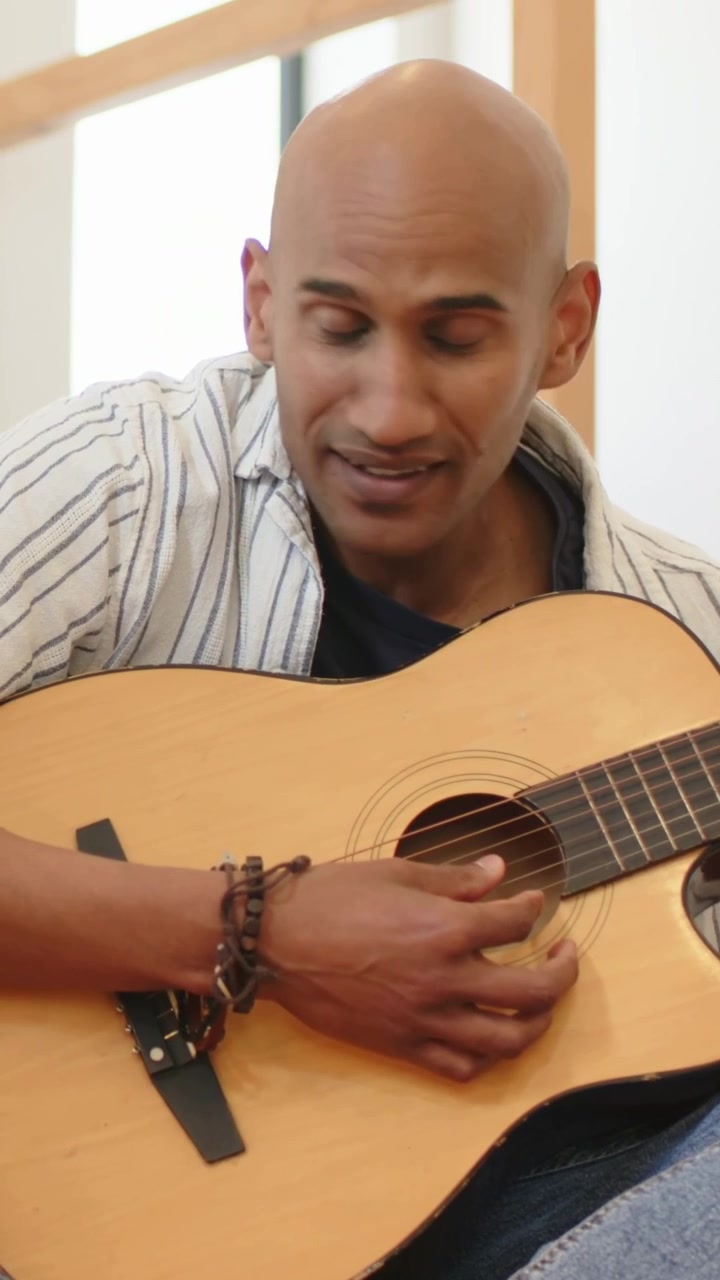 Video verticale: Primo piano di un uomo che inizia a sorridere verso un ascoltatore, strimpellando la chitarra a casa, con una maglietta a righe.