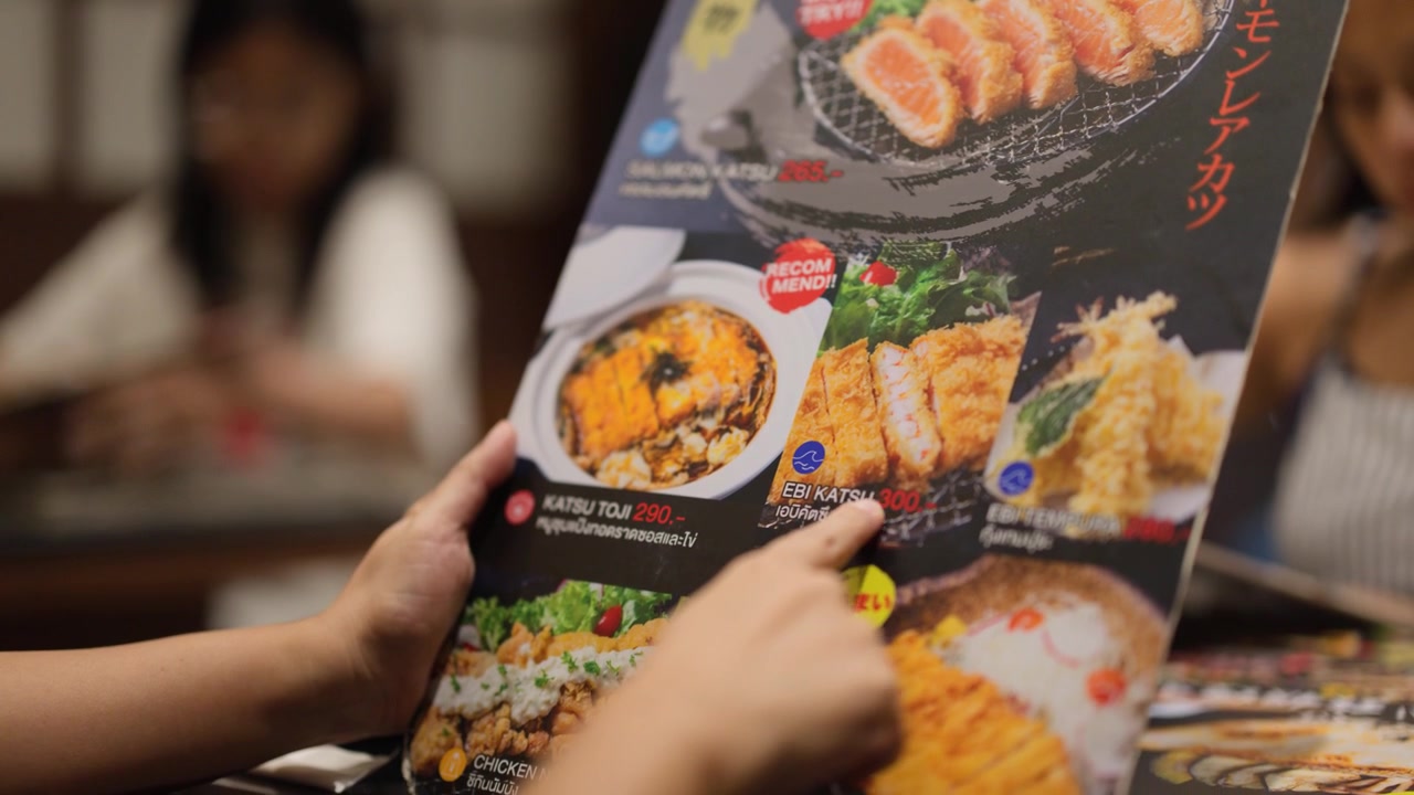 Detail rukou držících barevné menu při výběru jídla v rušné restauraci.