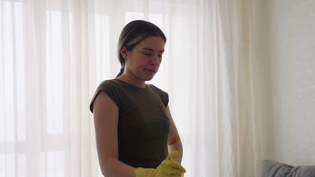 Cleaner in casual clothing wearing yellow gloves walks forward holding mop in bright living room with sunlight through curtains, partial view of sofa visible in background