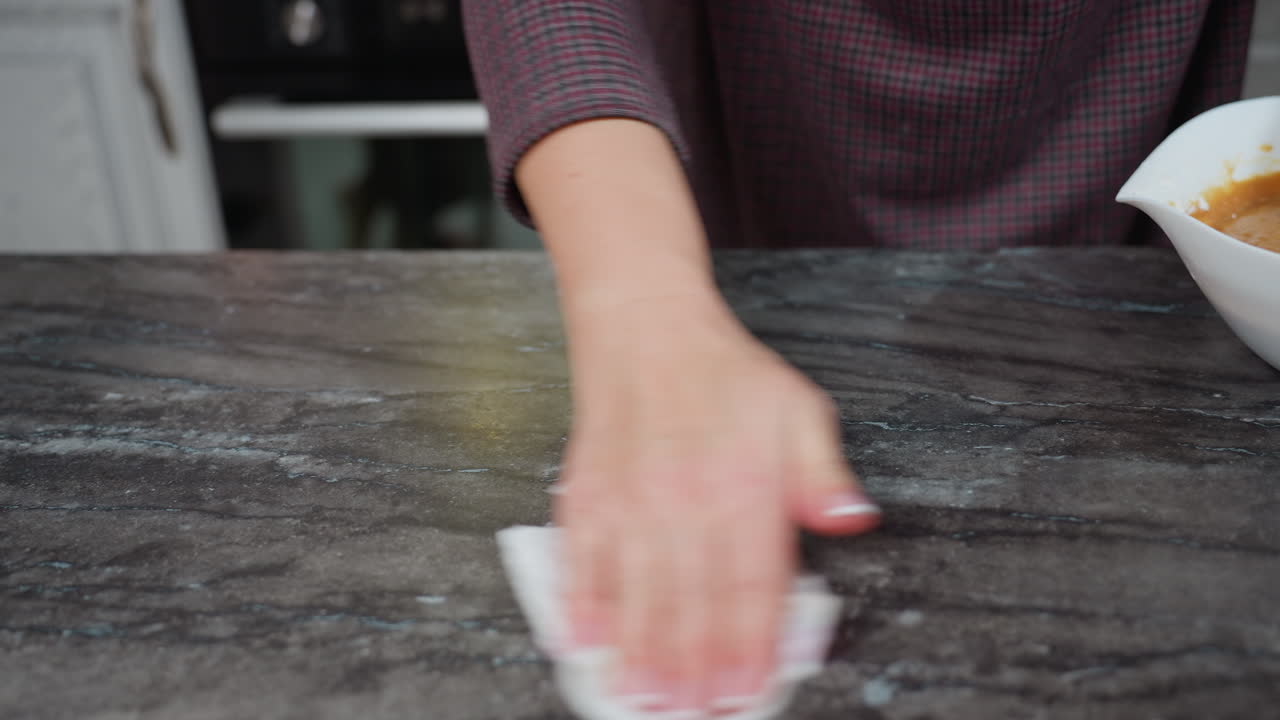 Visuale parziale di una donna che pulisce il piano di lavoro della cucina con un panno, assicurando la pulizia dopo aver cucinato. La superficie in marmo riflette la luce mentre la mano si muove.