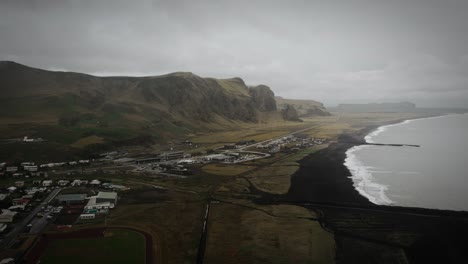 Aerial-black-sand-beach-and-town-vik,-ocean-scenery,-gren-grassy-mountains,-Iceland