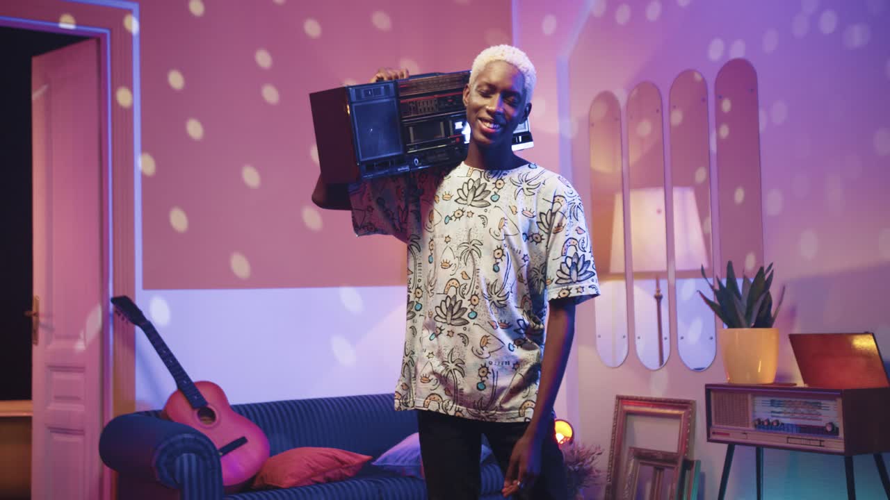 행복한 쾌활한 젊은 아프리카계 미국인 남자가 집에서 복고형 파티에서 거실에 서서 어깨에 복고풍 붐 박스를 들고 카메라를 보고 있습니다.