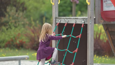 a little fair-haired girl climbs a jungle gym net and then takes a slide on the outdoor playground