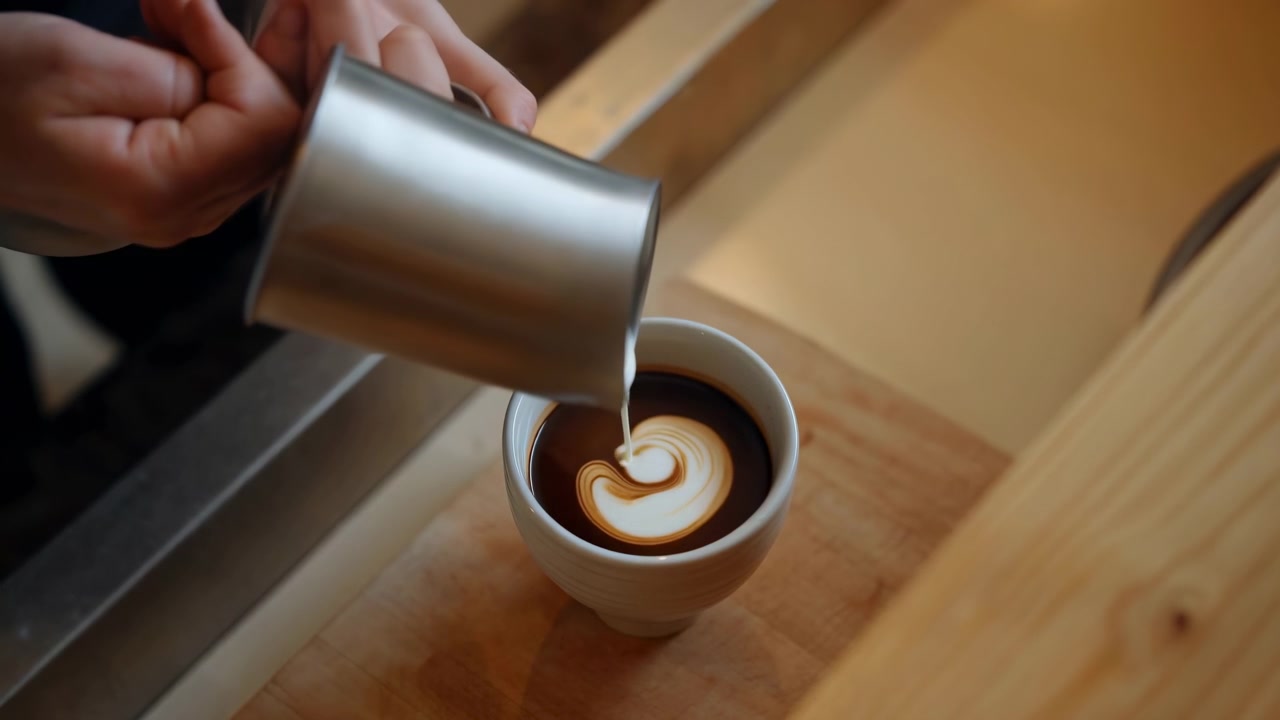 Nærbillede af en barista, der hælder dampet mælk fra en kande i rustfrit stål ned i en hvid keramisk kop sort kaffe, hvilket skaber en latte art roset med bløde koncentriske cremefarvede ringe på en varm køkkenbordplade.