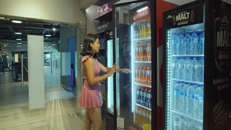 una chica joven en ropa deportiva abriendo y tomando una bebida del refrigerador en el gimnasio