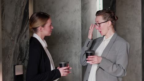 una chica rubia de mediana edad con gafas en un traje de negocios con café en las manos se comunica con su colega de negocios una joven chica rubio en una oficina moderna. diálogo entre dos colegas de negocios en la oficina