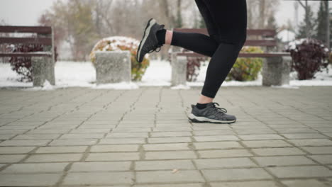 close-up leg view of lady jogging in black sneakers and leggings along snowy park pathway, surrounded by frosty winter scenery with benches, colorful bushes and distant resident