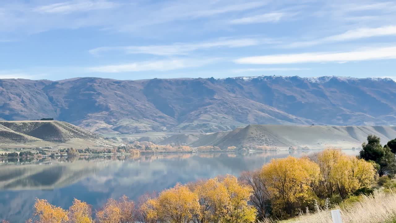A serene drive along Queenstown's lake with vibrant autumn foliage, clear skies, and majestic mountains in the background