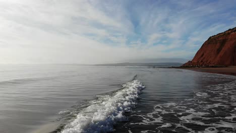 Low-Shot-Tracking-Entlang-Einer-Welle,-Die-Auf-Einen-Strand-Mit-Einem-Wunderschönen-Himmel-Und-Klippen-Dahinter-Rollt