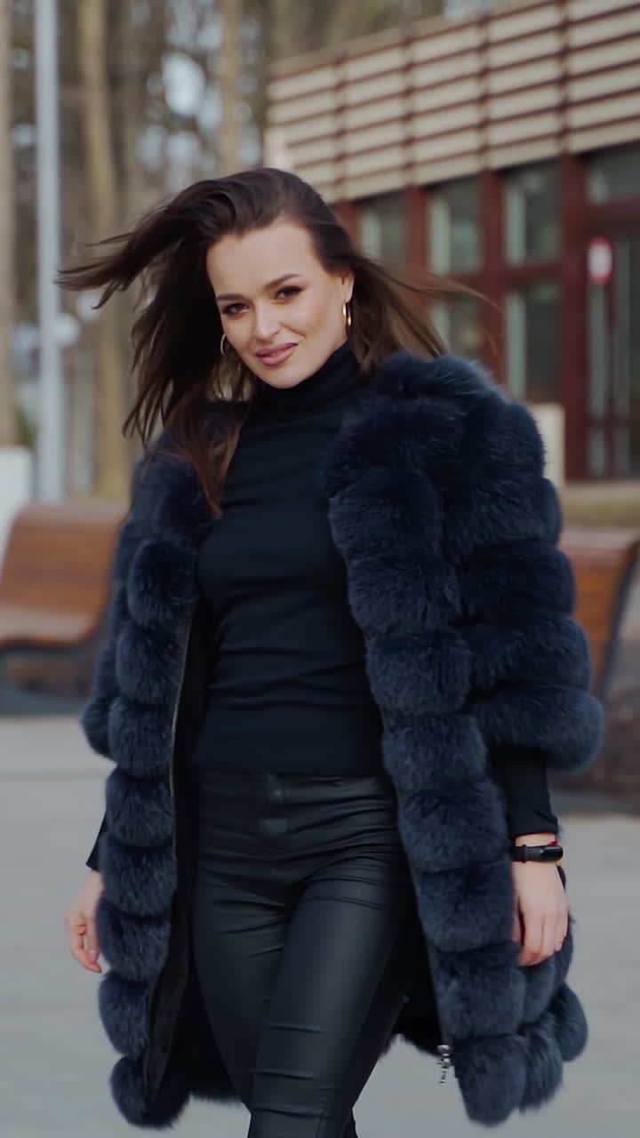 Attractive girl posing in the park. Beautiful young woman in black fur coat looking on camera and smiling outdoors. Slow motion. Vertical video