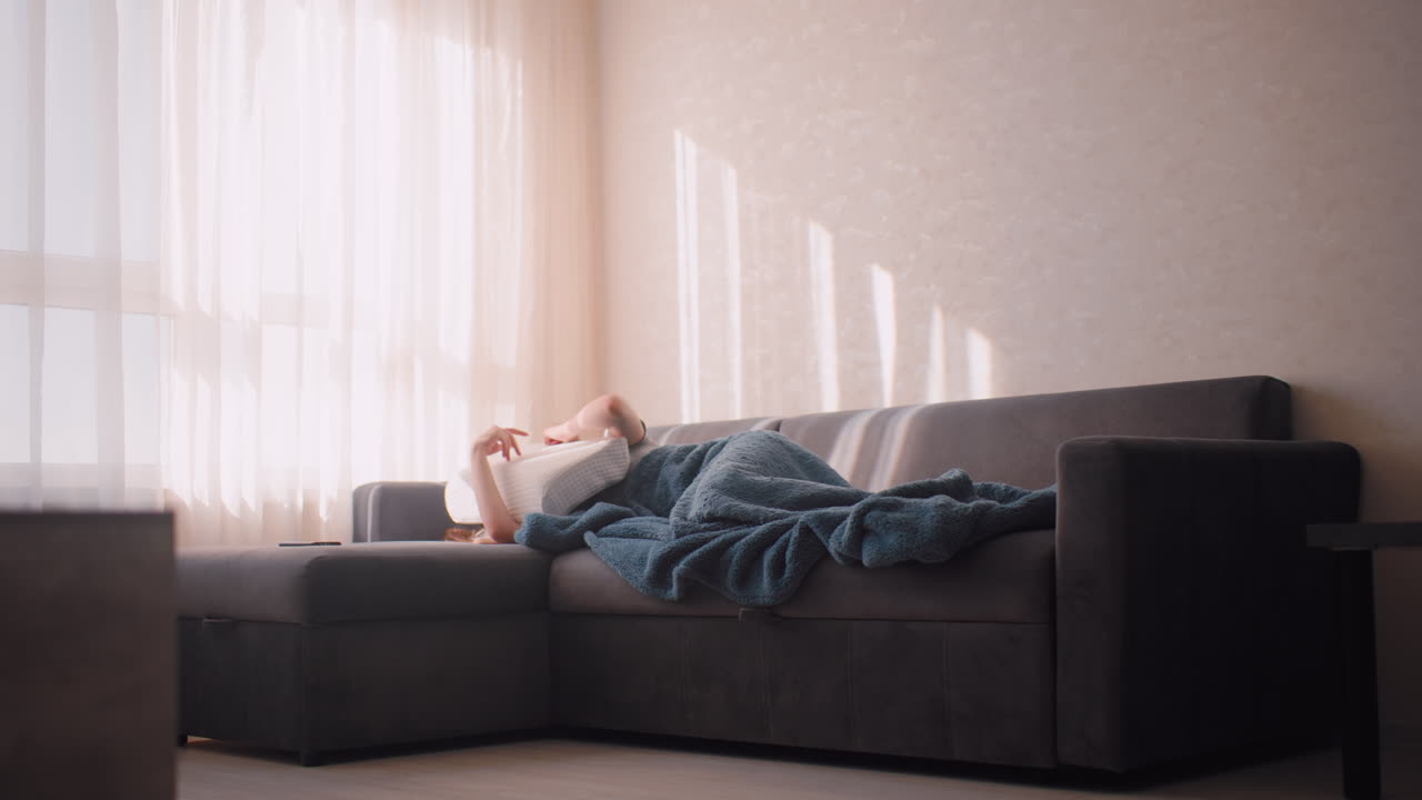 Woman sleeping on dark couch uses pillow to shield face from harsh sunlight streaming through curtains, blue blanket covering body, morning rays casting shadow on wall with nearby side stool in view