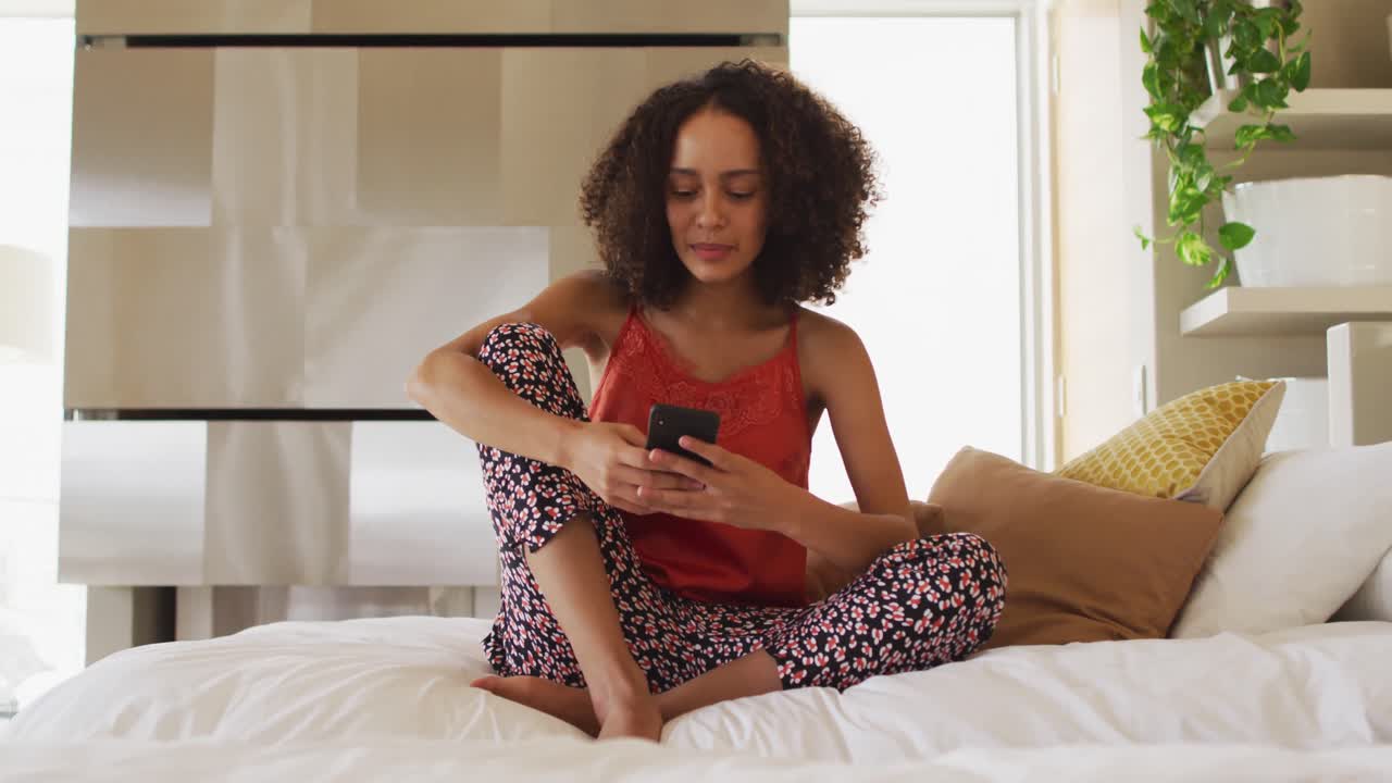 mujer afroamericana sentada en la cama usando un teléfono inteligente sonriendo