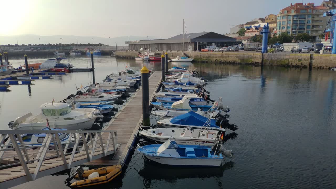 Un pescatore prepara gli attrezzi tra le barche da pesca ormeggiate nella darsena del porto con accesso tramite rampa alla piattaforma, in una tranquilla e soleggiata mattina d'estate, inquadratura fissa.