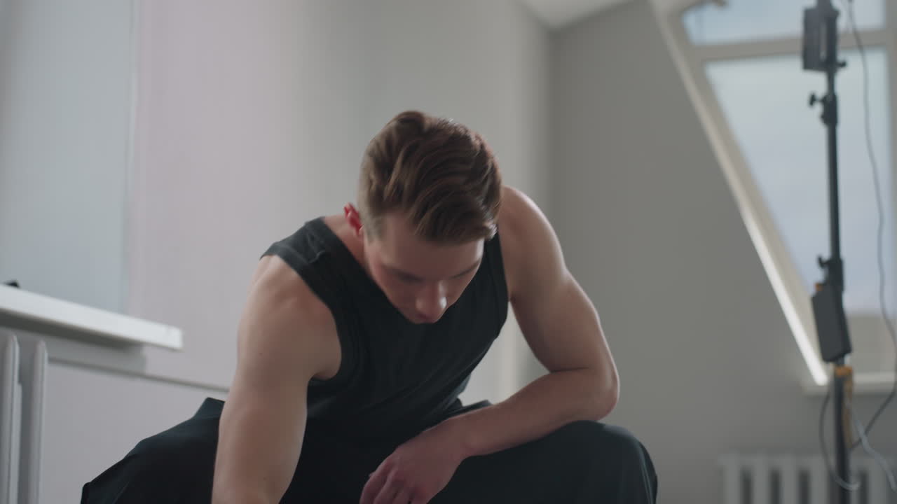 Young man in black tank top sits on stool by open window, adjusts black jacket on lap as sunlight filters through high rise building outside in minimalist modern apartment interior