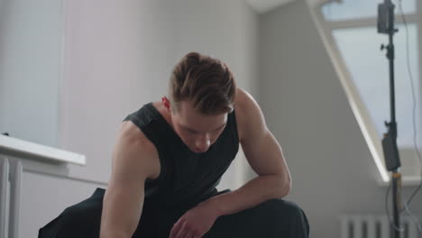 young man in black tank top sits on stool by open window, adjusts black jacket on lap as sunlight filters through high rise building outside in minimalist modern apartment interior