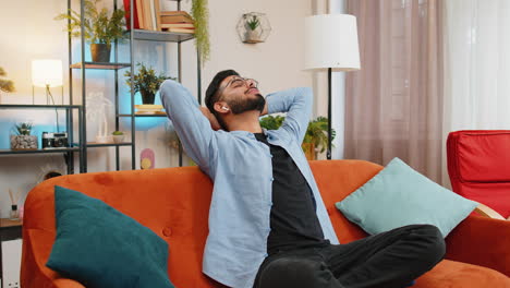 happy relaxed hispanic man resting on home couch with wireless earphones listening ambient music