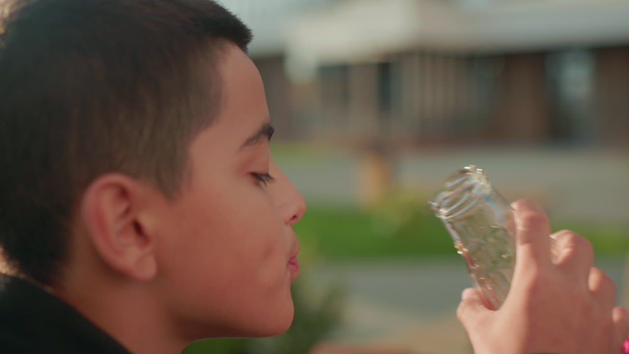 Close up side view of kid drinking fruit juice from glass bottle outdoors, holding it with both hands, enjoying refreshing beverage under natural sunlight with relaxed expression in urban environment