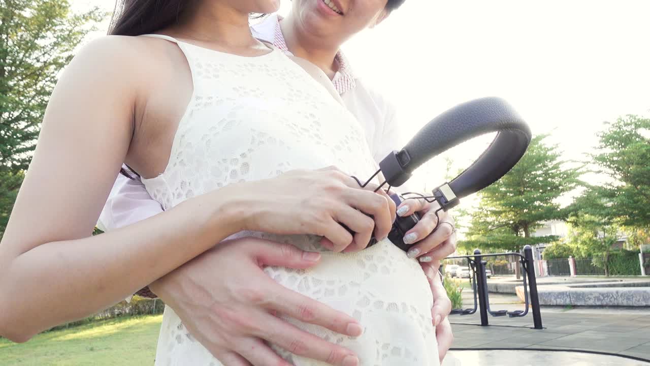 행복한 임신 아시안 여성과 남편이 정원에서 배에 헤드폰을 달고 있습니다.