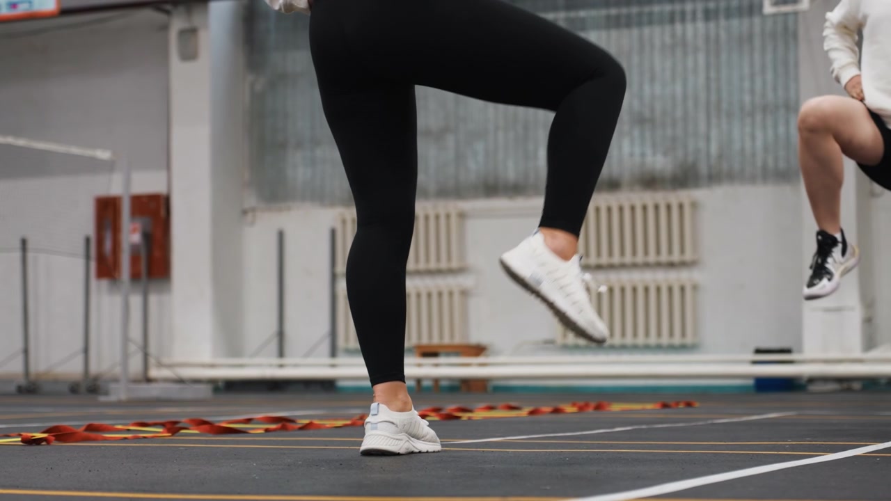 Indoor badminton court feet performing partner footwork drill, two players in athletic wear execute quick lateral steps along white lines, sneakers gripping rubber surface, coachlike repetition builds