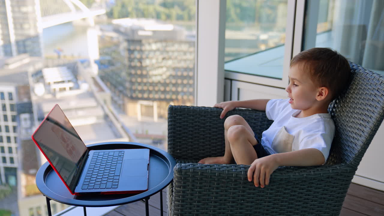 Calm relaxed baby boy sits leaned back in the armchair near the panoramic window. Little boy watches video on laptop