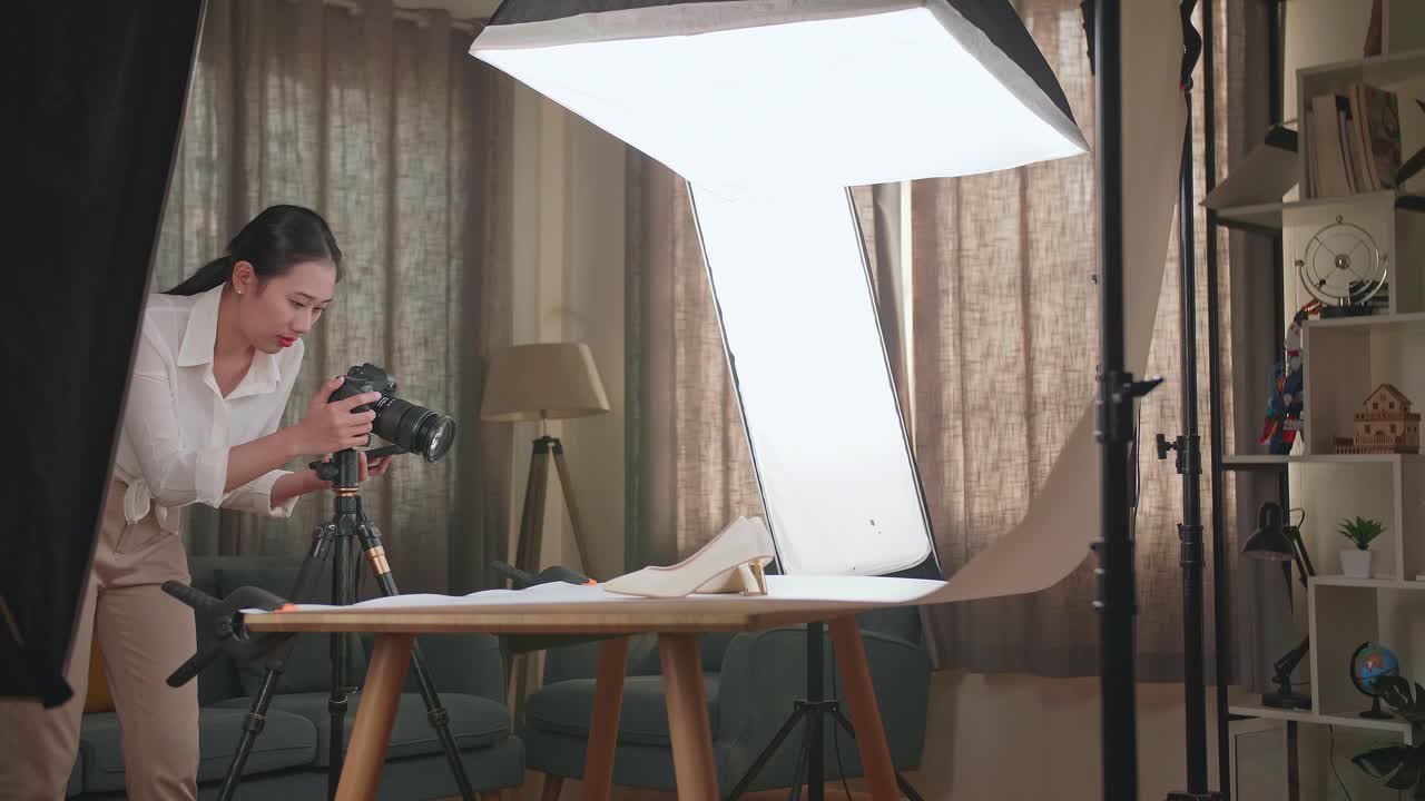fotógrafa asiática hablando en su teléfono inteligente mientras toma fotos de zapatos femeninos en un estudio casero