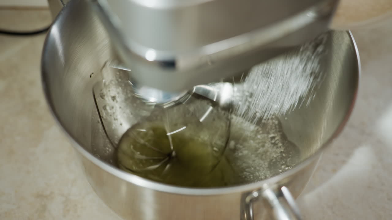 Cook using stand mixer to combine egg whites and flour in stainless bowl while pouring sugar, showcasing smooth blending process during dessert preparation in bright kitchen