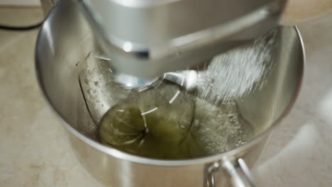 cook using stand mixer to combine egg whites and flour in stainless bowl while pouring sugar, showcasing smooth blending process during dessert preparation in bright kitchen