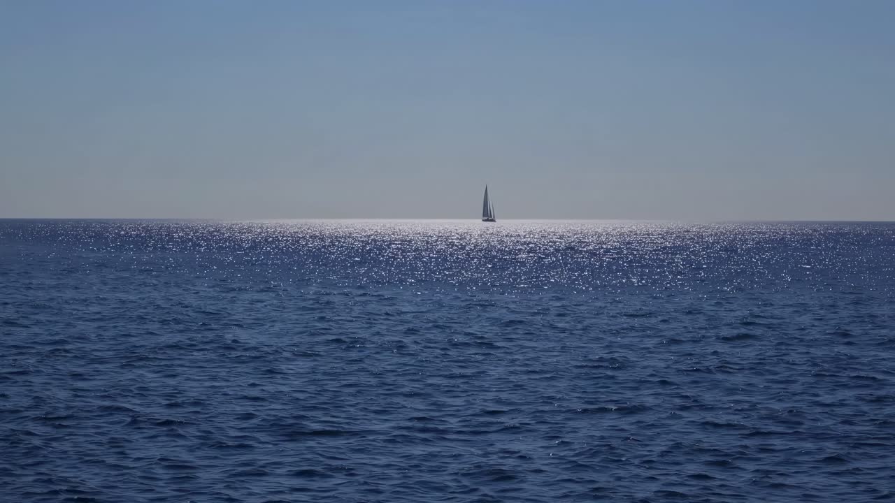 A serene video of a distant sailboat on a shimmering sea, captured from a low-angle perspective