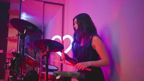 drummer girl playing electronic drums under pink and purple lighting in cozy indoor studio, heart-shaped neon light and mirror in background reflecting part of room and glowing drum equipment