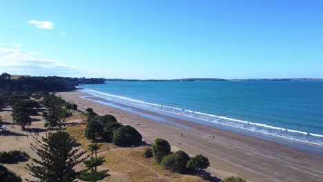 drone shot, flying over trees towards a beautiful beach on a sunny day in long bay, new zealand