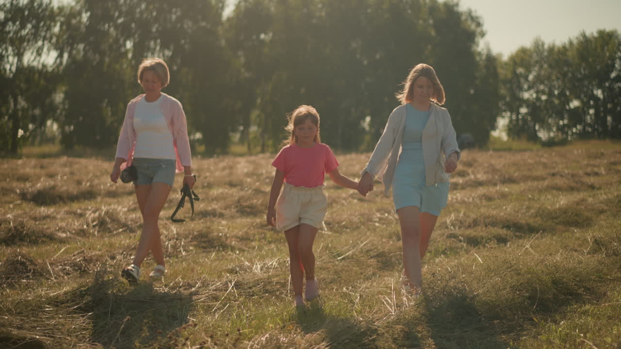 두 여동생은 은 날 농장에서 아이와 함께 걸어가고, 머리카락과 옷이 바람에 날아다니며, 아이가 이모의 손을 잡고 산책하면서, 은 공기와 자연을 즐기고, 초록색 나무가 배경입니다.