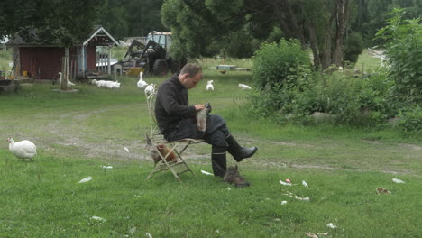 man greasing his leather hiking boots on a farm, boy cleaning his shoes on a chair, chicken and geese in the background, finland