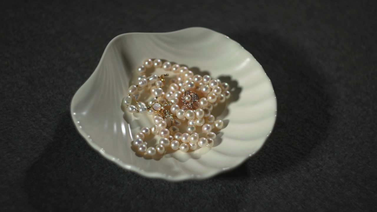 Fili di perle in ciotola di ceramica, mano di gioielliere solleva collana, ombra morbida, sfondo scuro testurizzato, caldo