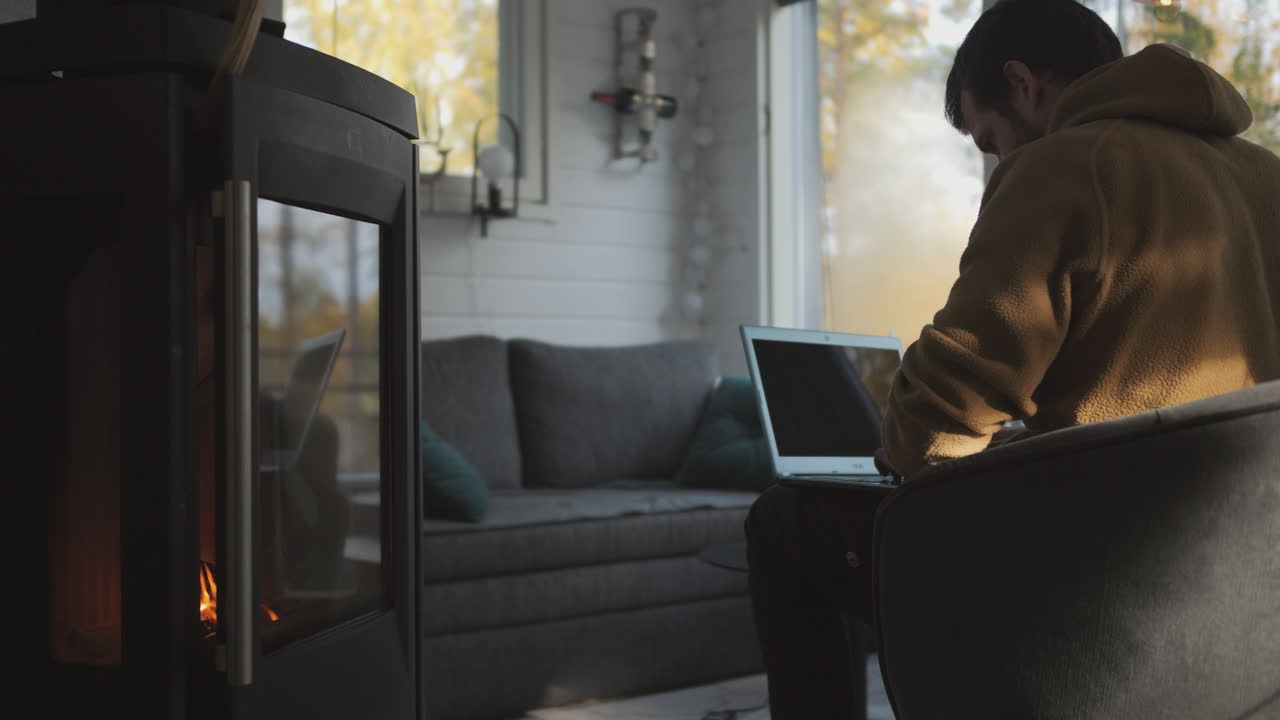 Man works on laptop in front of fireplace in cozy small cabin home