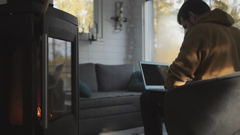 man works on laptop in front of fireplace in cozy small cabin home