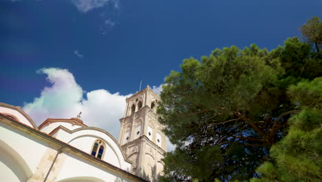 Vista-En-ángulo-Bajo-De-La-Icónica-Torre-De-La-Iglesia-En-Lefkara,-Chipre,-Con-Su-Arquitectura-Distintiva-Enmarcada-Por-Un-Pino-Verde-Contra-Un-Cielo-Azul-Parcialmente-Nublado