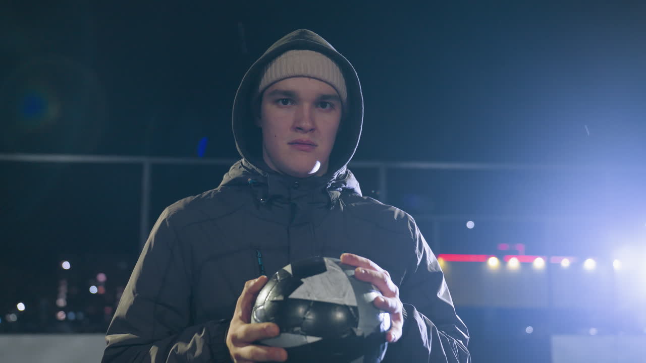 niño joven con capucha sosteniendo una pelota, mirando triste bajo la luz brillante de la calle durante la noche, reflejando la lucha emocional y el pensamiento profundo